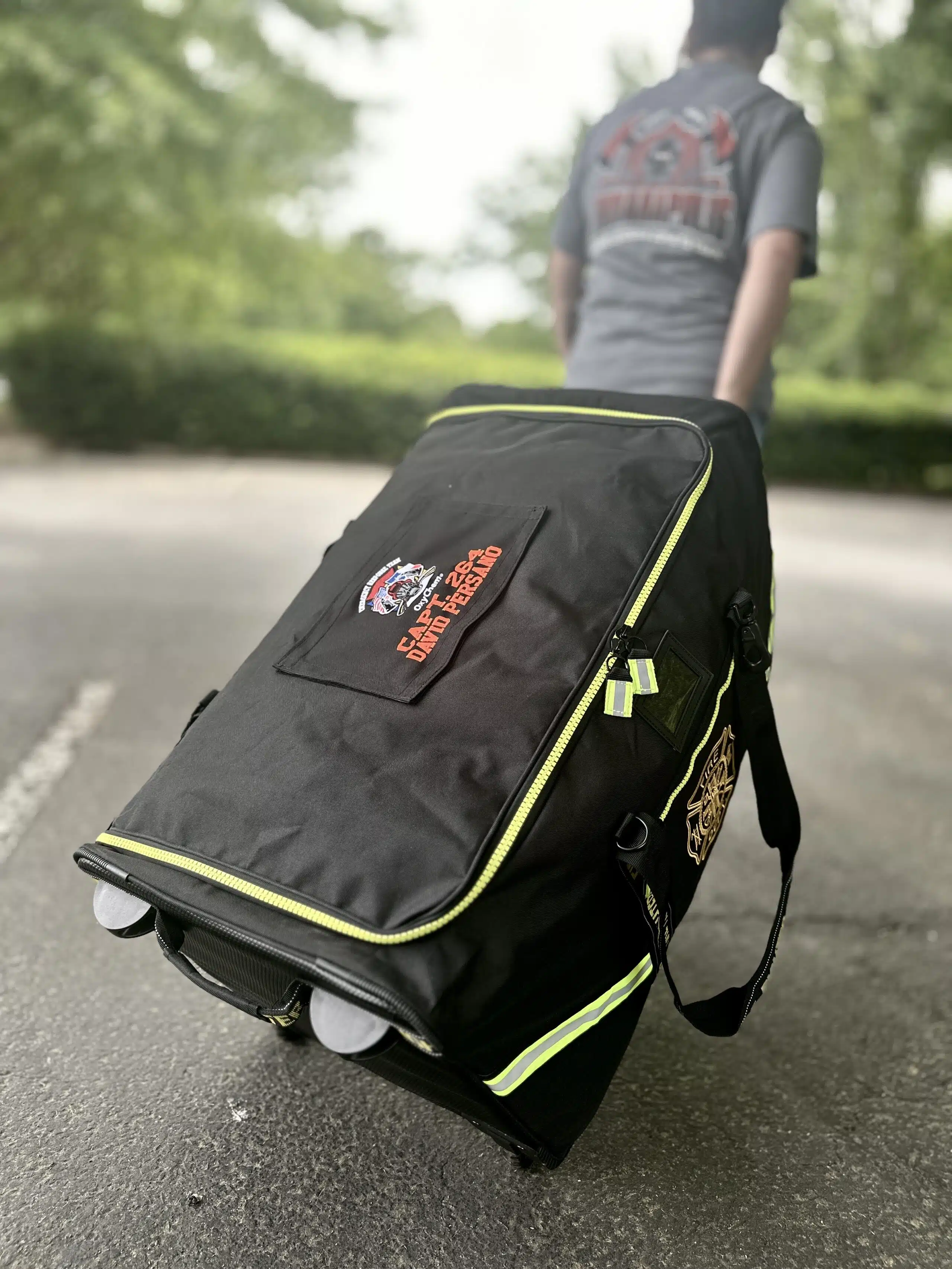 Person pulling a large black rolling gear bag behind them on asphalt. Bag top flap has matching embroidered patch.
