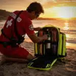 Beach lifeguard opening a fluorescent-yellow medical backpack with an oxygen cylinder inside at sunset