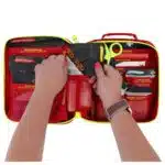Hands lifting a color-coded pouch from an open red soft-shell first aid kit filled with labeled supplies